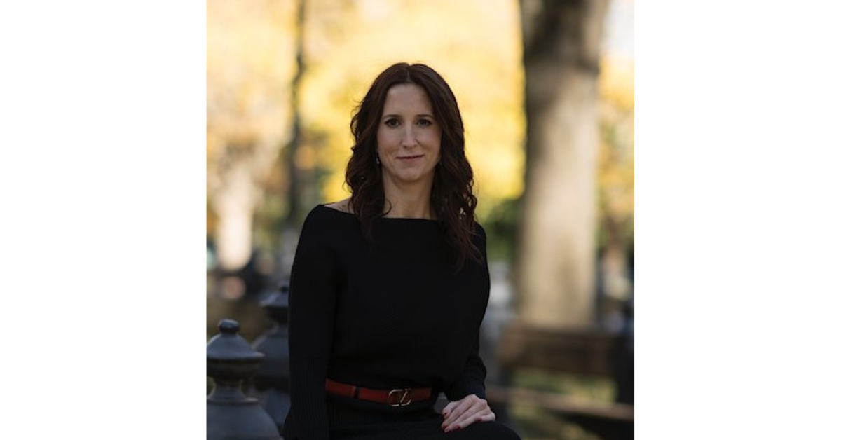 Tracy Ongena seated outdoors in a black dress with autumn trees in the background
